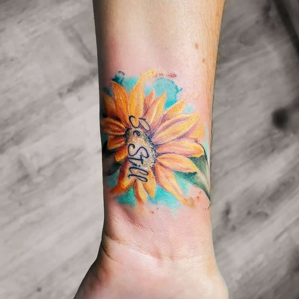 Sunflower tattoo on the wrist with the text "Be Still" written in cursive on the petals