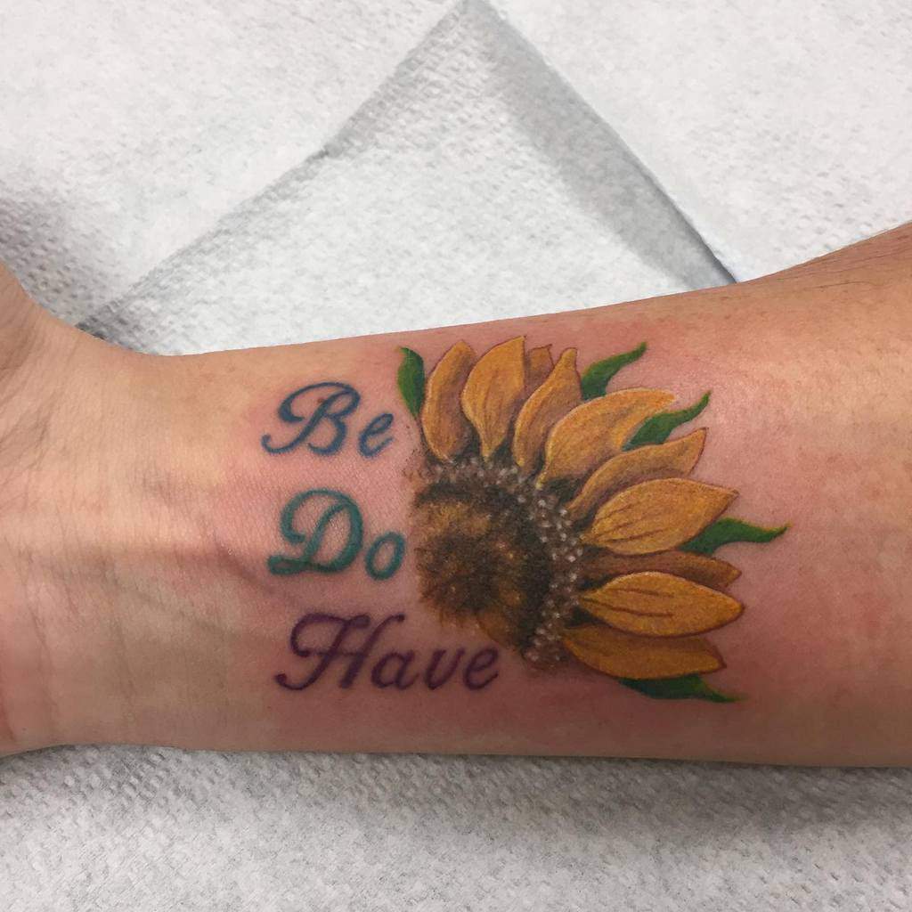 Tattoo of a sunflower with the words "Be Do Have" on a wrist