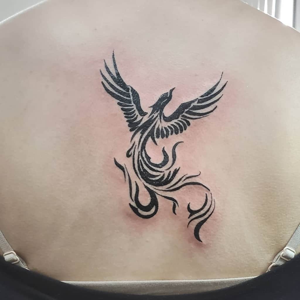 A tribal tattoo of a stylized black phoenix with spread wings graces the person's upper back