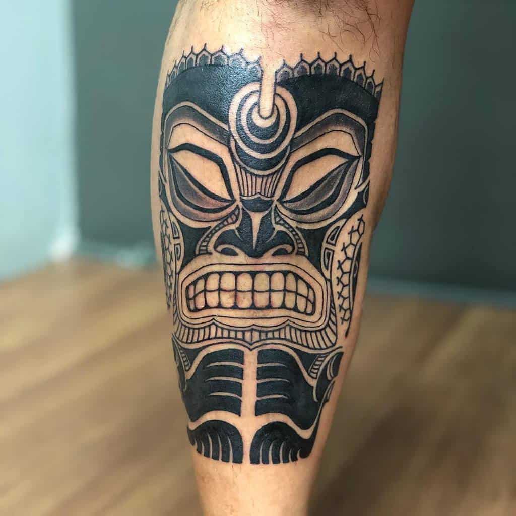 A striking tattoo of a fierce tribal mask adorns the person's calf, intricately detailed with bold black lines