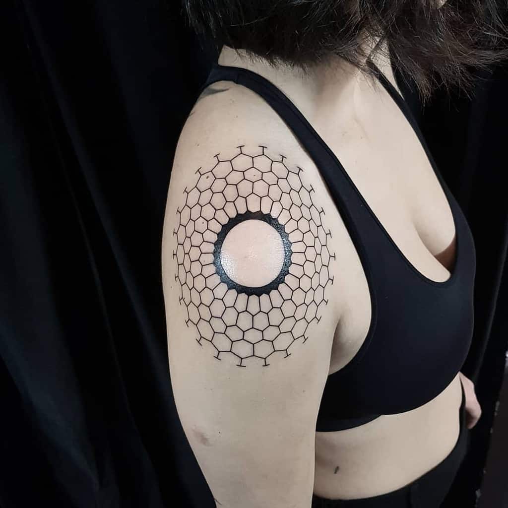 The tattoo features a geometric design on the shoulder, with a central circular shape surrounded by a honeycomb pattern, creating a striking and symmetrical look with bold black lines