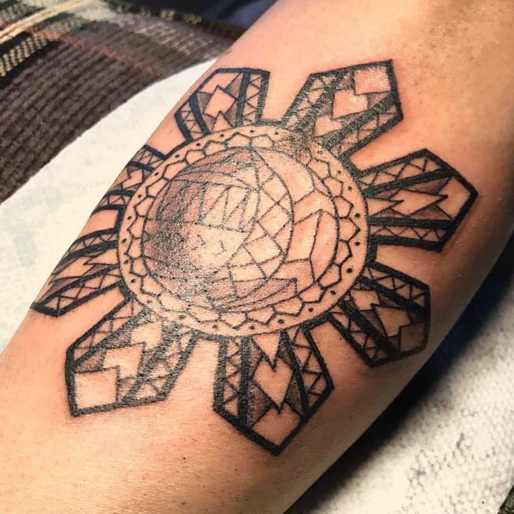 The tattoo features a geometric sun design on the forearm, with intricate patterns and sharp angles extending from a central circular shape, creating a bold, symmetrical look