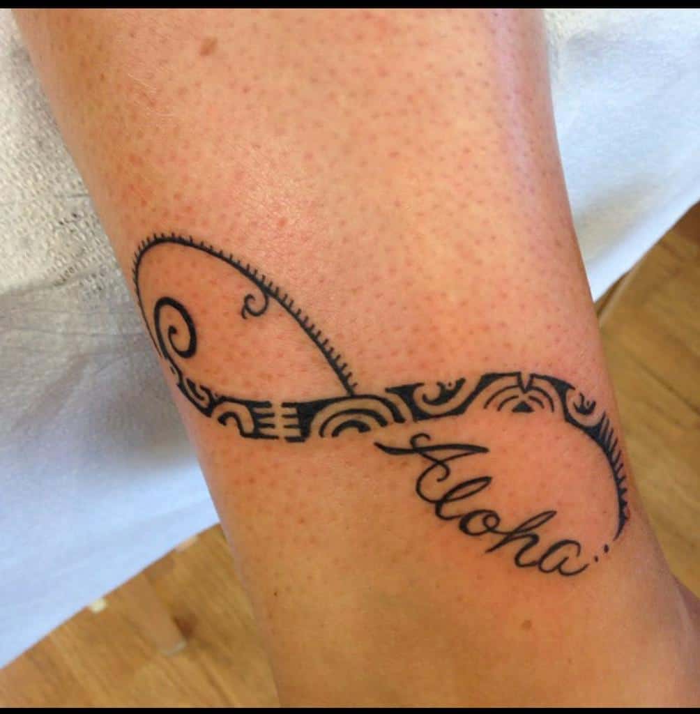 Tattoo of an infinity symbol with tribal patterns and the word "Aloha" on a leg
