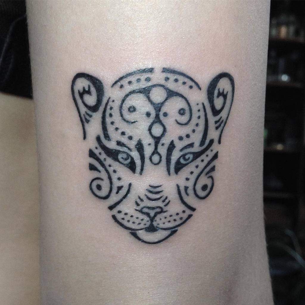Black tribal-style tattoo of a stylized tiger face on a forearm, featuring intricate swirls and patterns