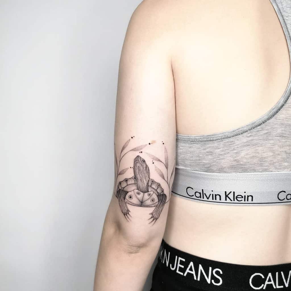 A detailed black ink tattoo of a turtle surrounded by leaves, placed on the upper arm near the shoulder