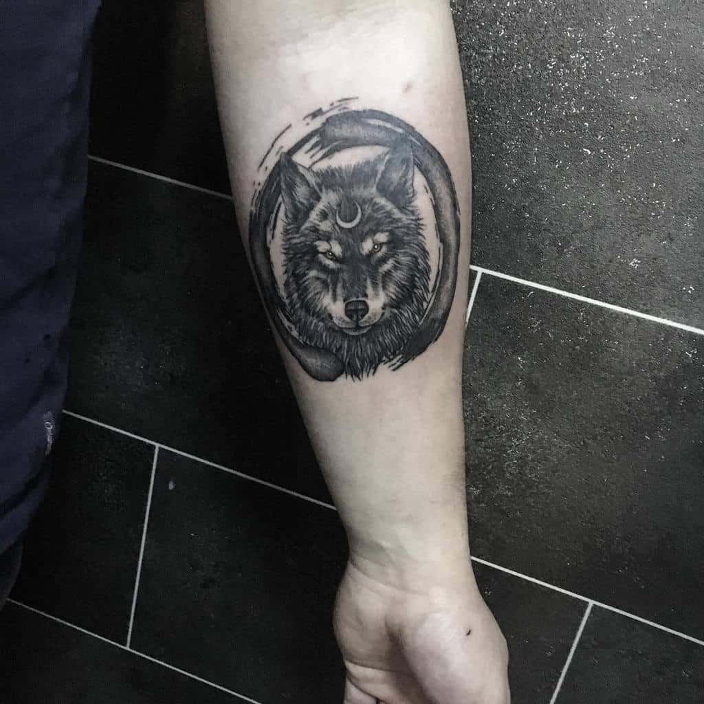 Detailed wolf head tattoo on the forearm, framed by a circular brushstroke and featuring a crescent moon symbol