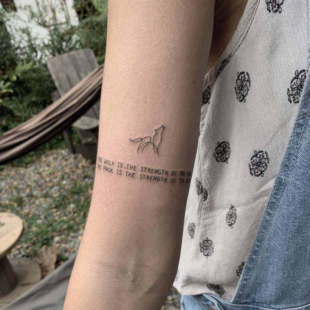 Small upper arm tattoo featuring a minimalist outline of a wolf with accompanying text that reads 'The wolf is the strength of the pack. The pack is the strength of the wolf.'