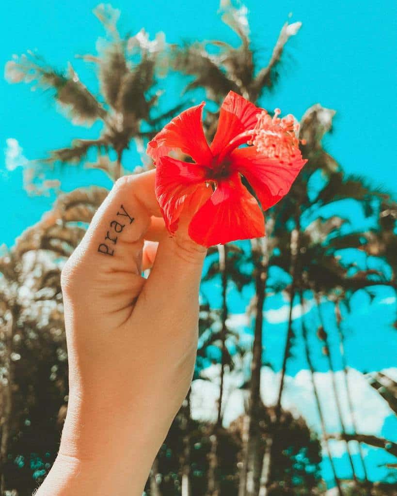 Minimalistic 'pray' tattoo on the side of a finger, paired with a vibrant red flower against a tropical palm tree backdrop