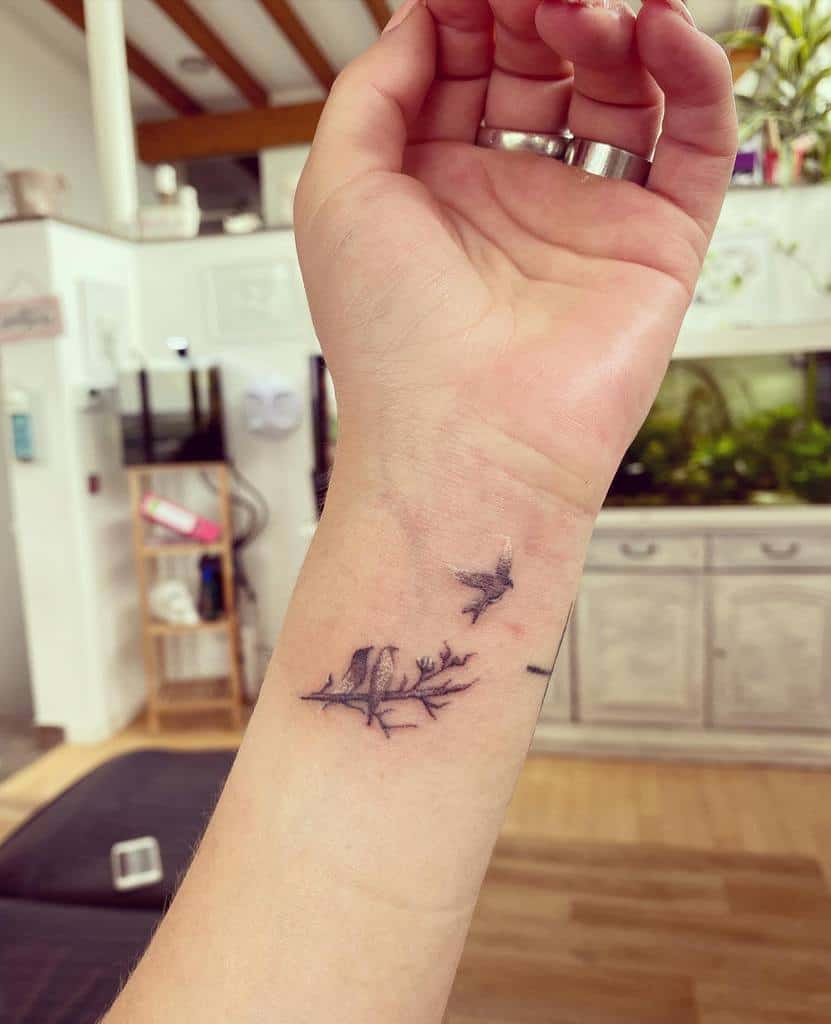 Delicate wrist tattoo featuring two birds perched on a branch and one in flight, symbolizing freedom, connection, and new beginnings