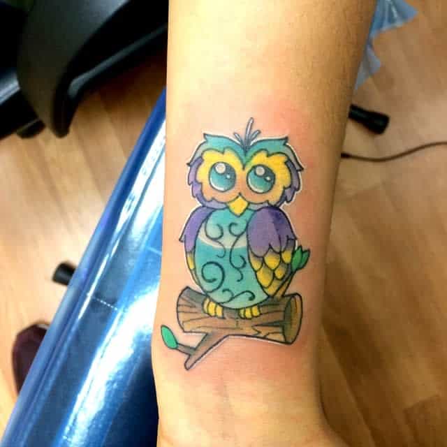 Colorful cartoon owl tattoo on a wrist, perched on a branch with a leafy background
