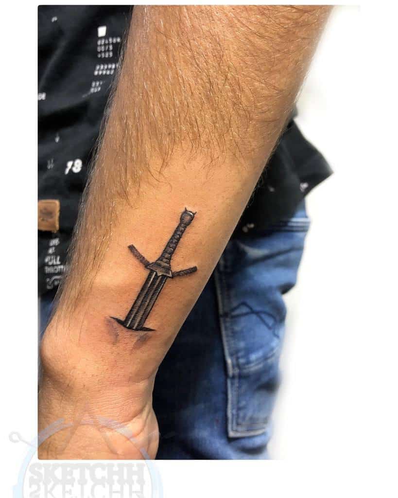 Wrist tattoo of a sword with a detailed hilt and blade, appearing to pierce the skin
