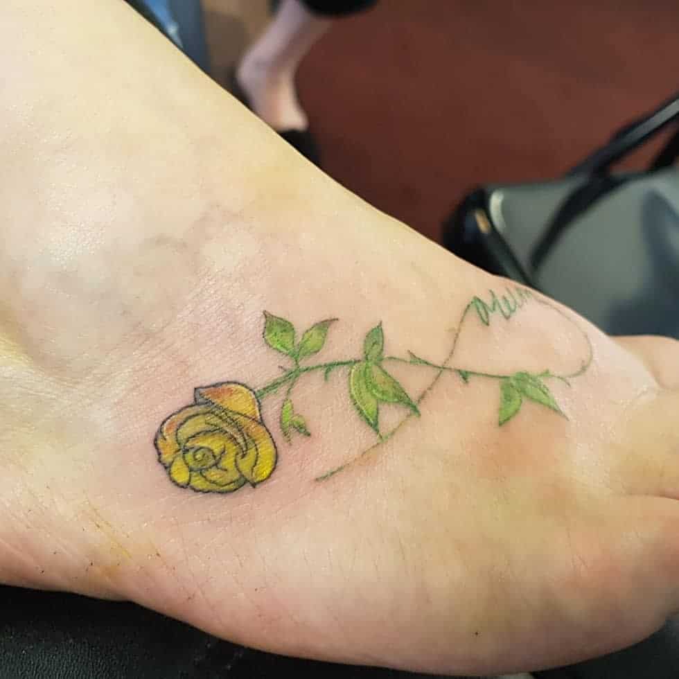 A yellow rose tattoo with green leaves on the top of a foot