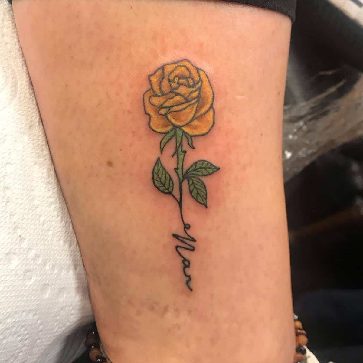 Tattoo of a yellow rose with green leaves and a stem that morphs into cursive text on an arm