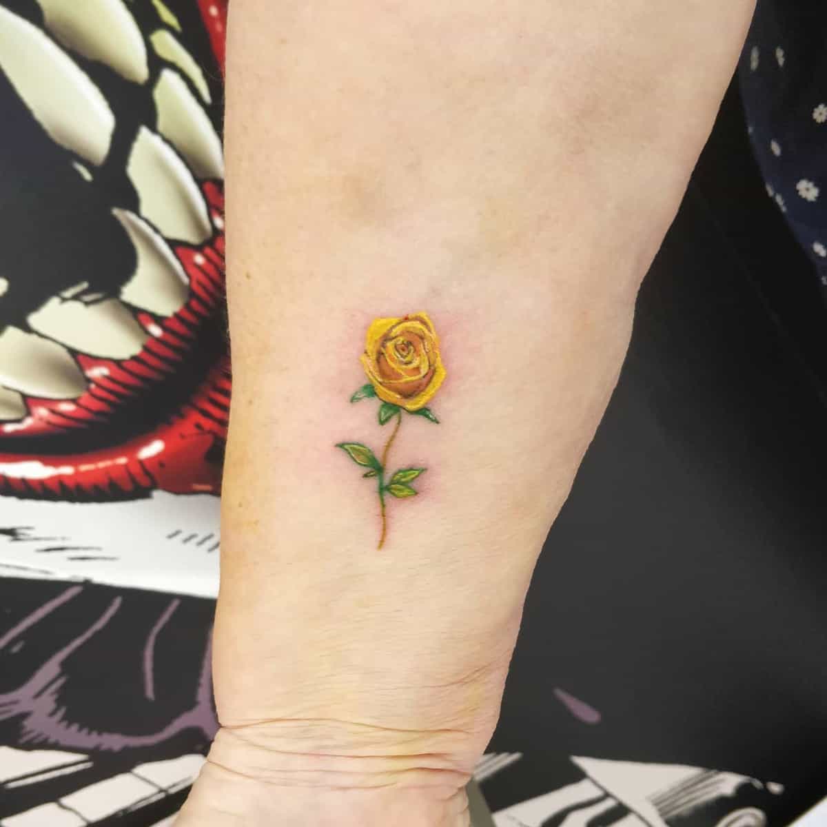 A small, detailed yellow rose tattoo on a forearm with green leaves and a stem