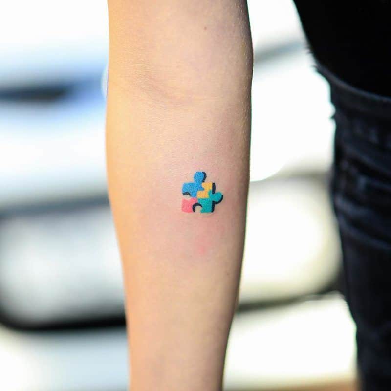 Small full color forearm tattoo of multi-colored puzzle piece with black drop shadow.