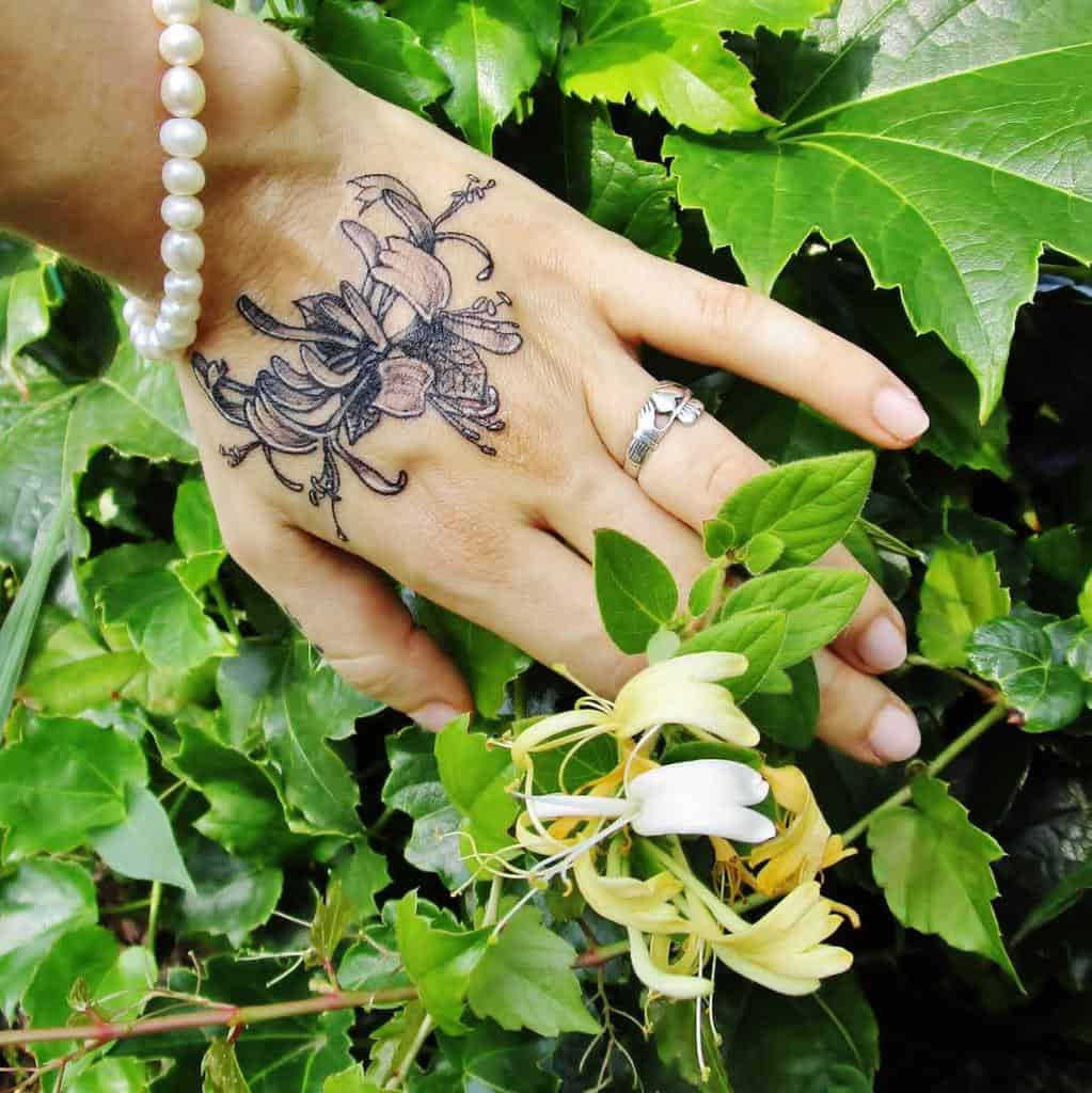 Small honeysuckle tattoo on the hand showcases delicate black and grey floral detailing, with elegant vines and flowers spreading naturally across the hand