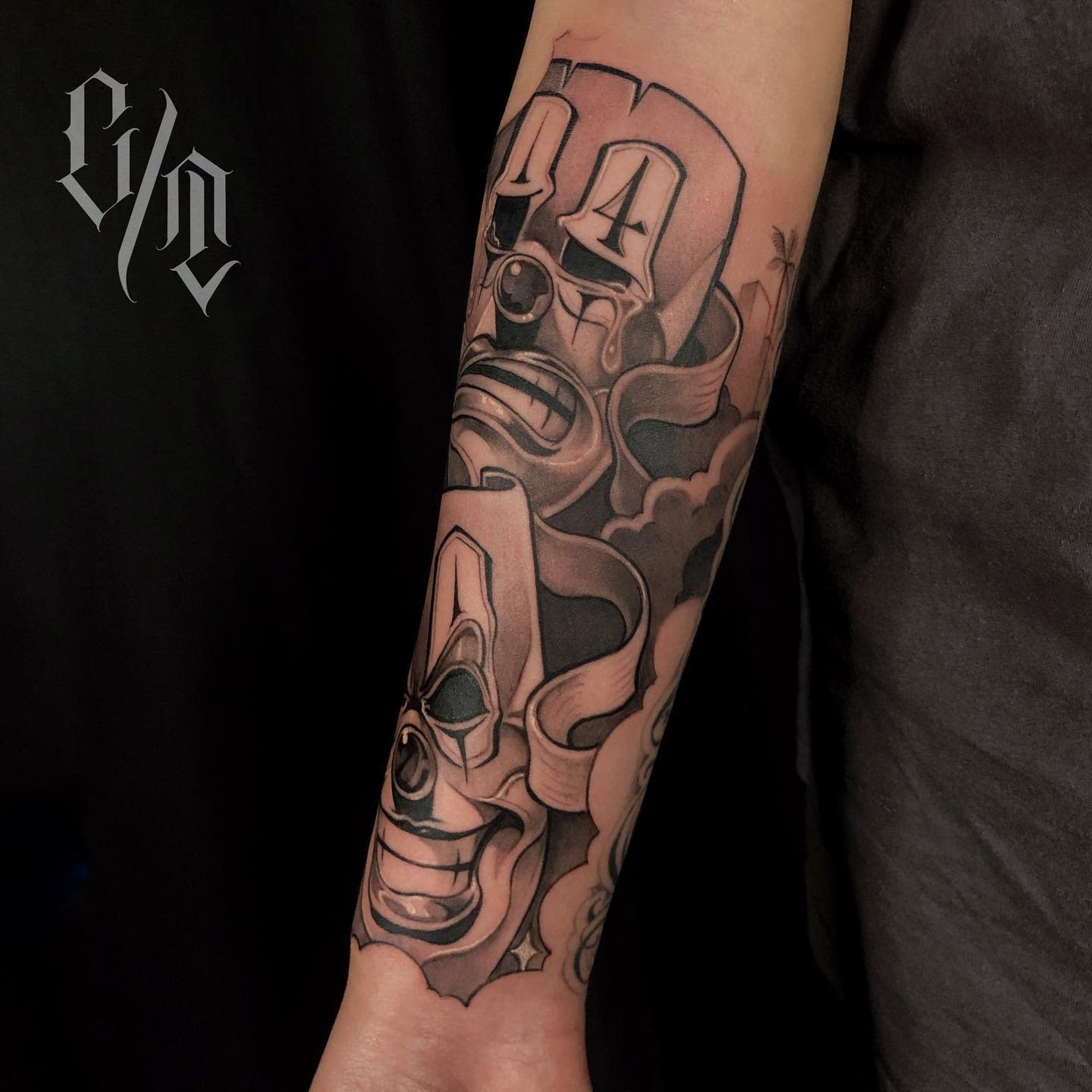 Forearm tattoo of two clown faces with detailed shaded patterns and letters: one is crying, the other laughing