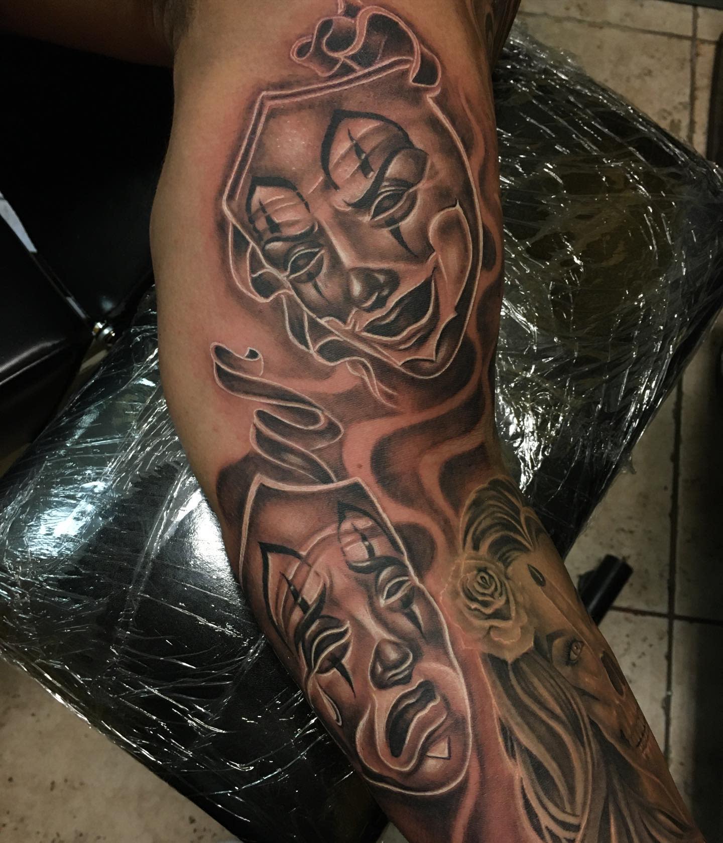 Upper arm tattoo of drama masks, one smiling and one crying, with intricate shading on the skin