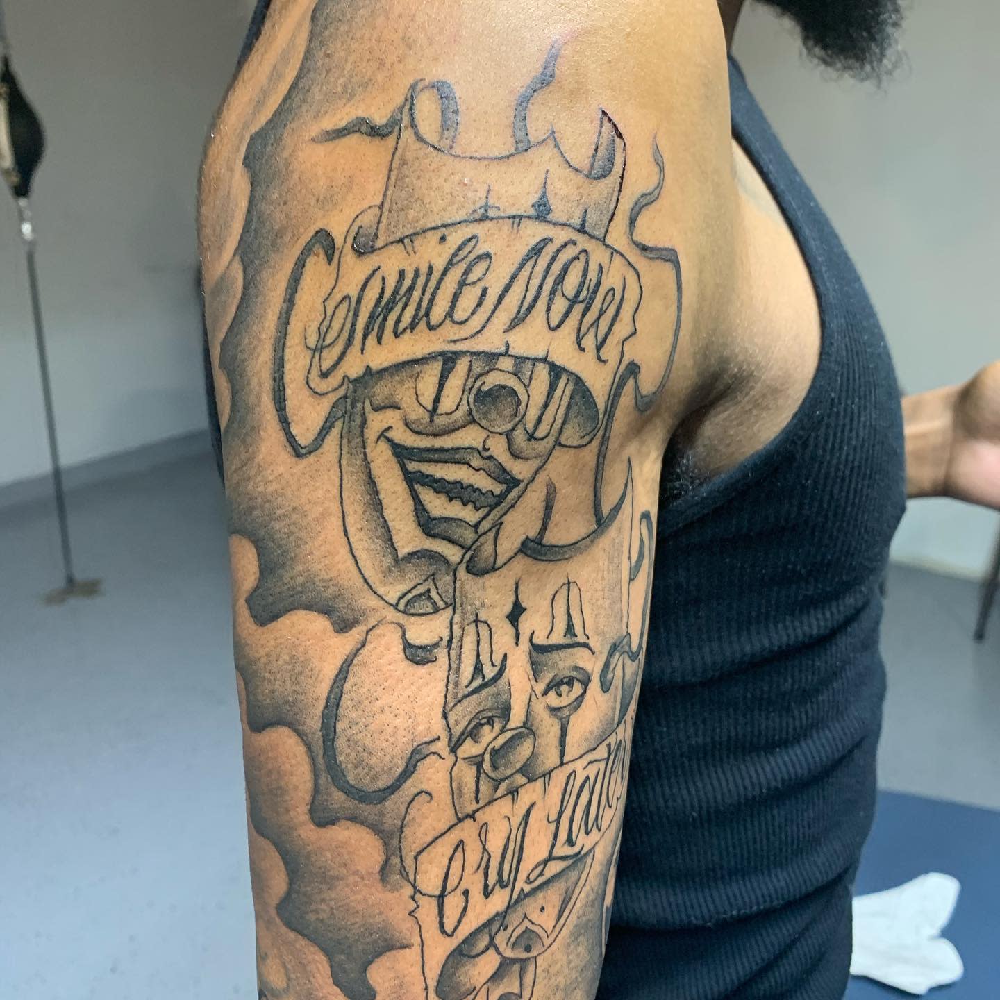 Black and gray tattoo of comedy and tragedy masks with "Smile Now, Cry Later" text on upper arm