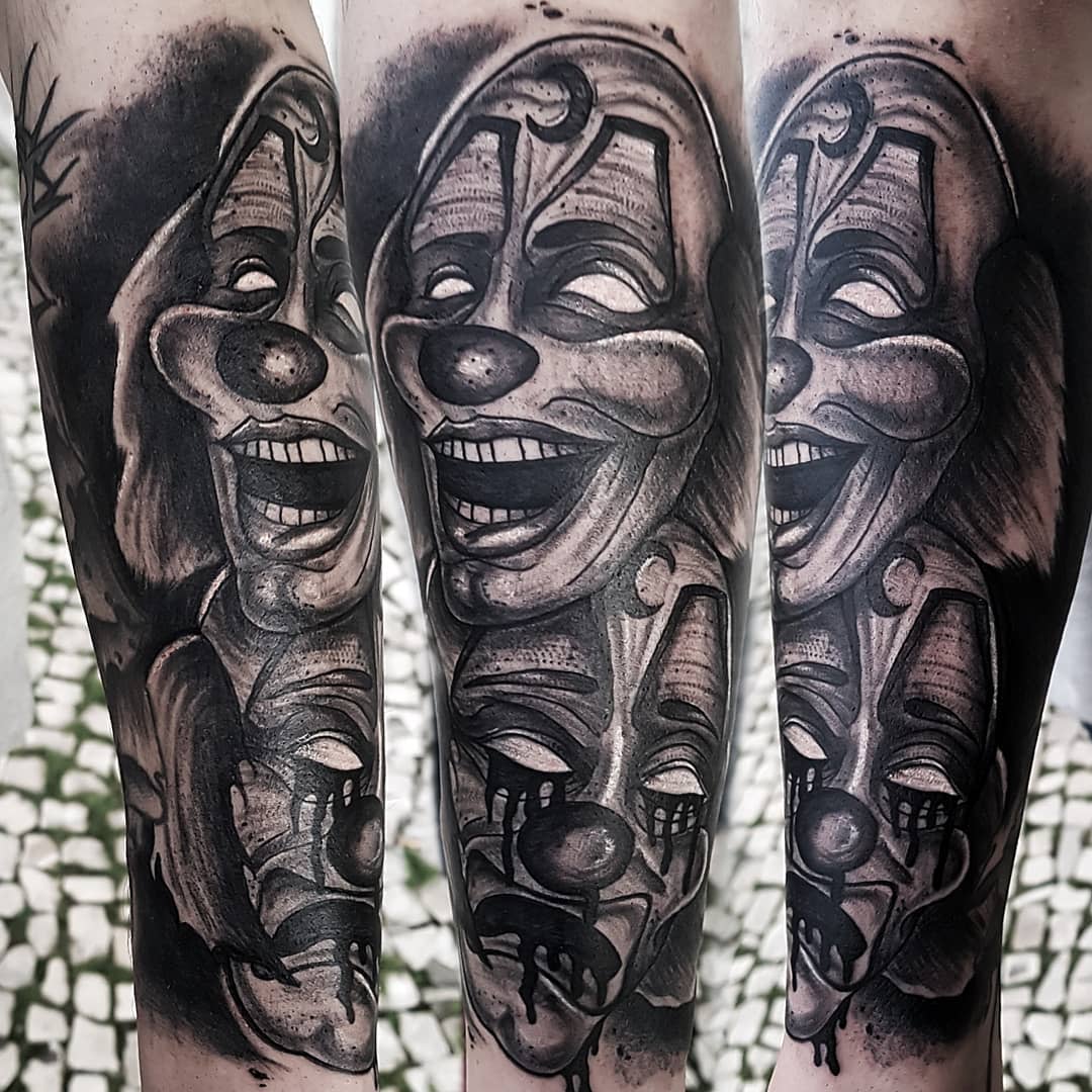 Black and grey tattoo of laughing, crying clown faces with dramatic shading on an arm