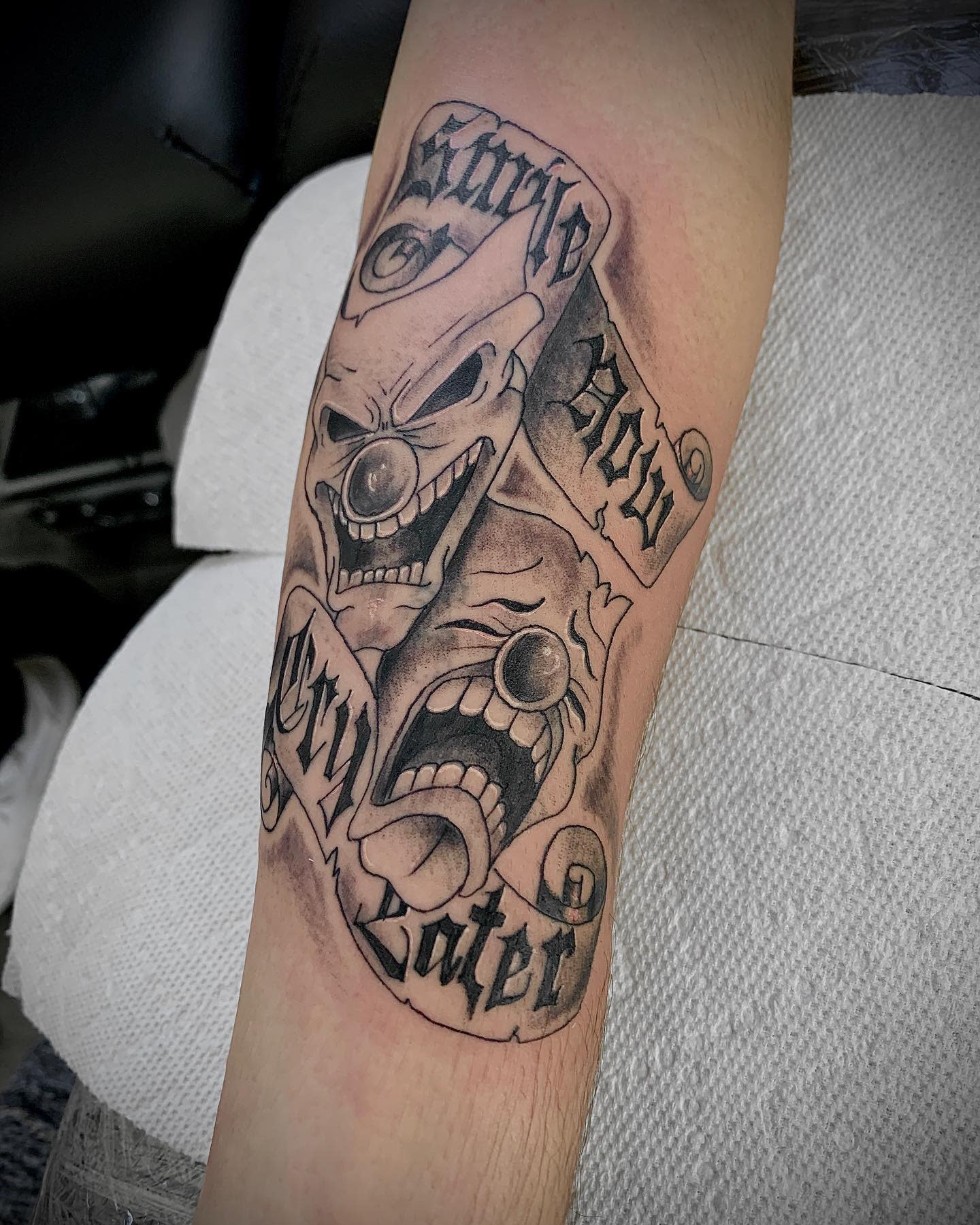Tattoo featuring two clown faces with a "Smile Now, Cry Later" text banner on an inner forearm