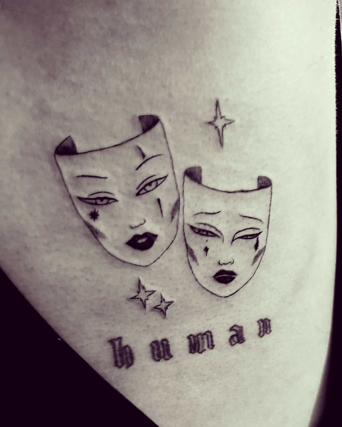 Tattoo of two masks with various facial expressions, stars around, and the word “human” below