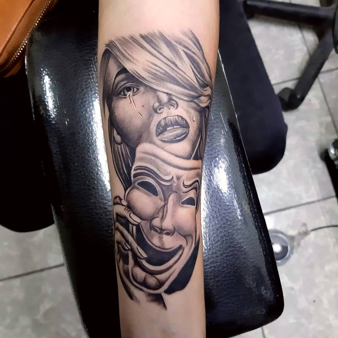Forearm tattoo of a sad woman with makeup and a hand holding a laughing mask over her face