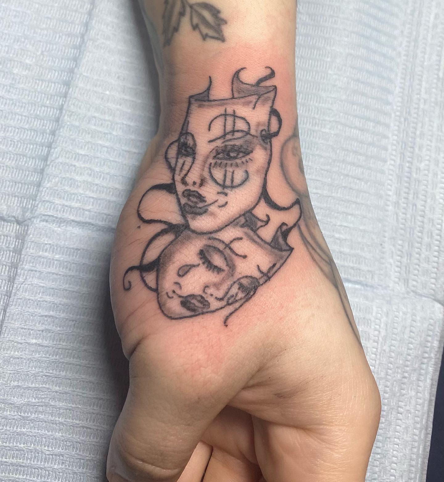 Tattoo with two masks featuring abstract faces and swirling lines on the back of a hand