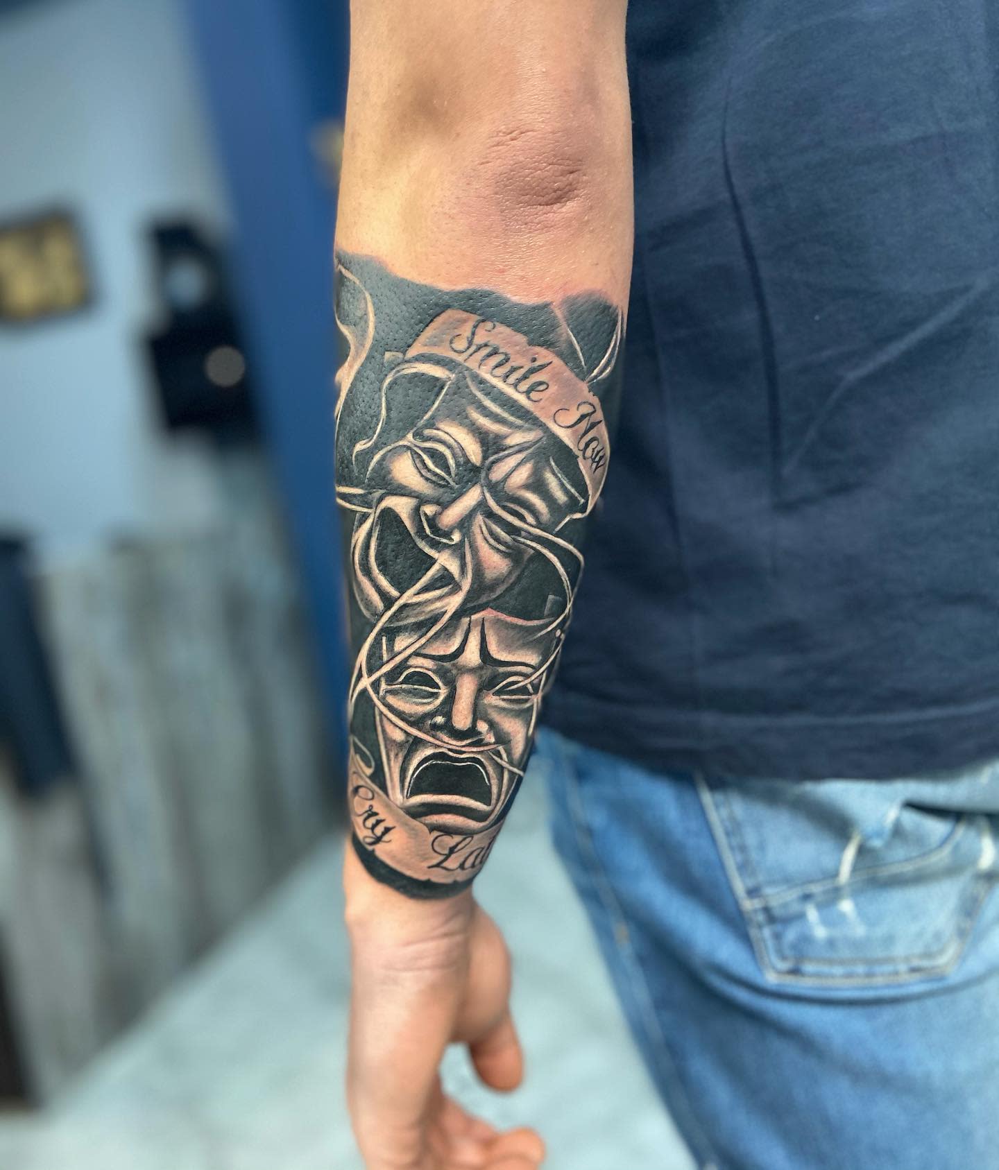 Forearm tattoo of theatrical masks in black ink; one smiling and one frowning