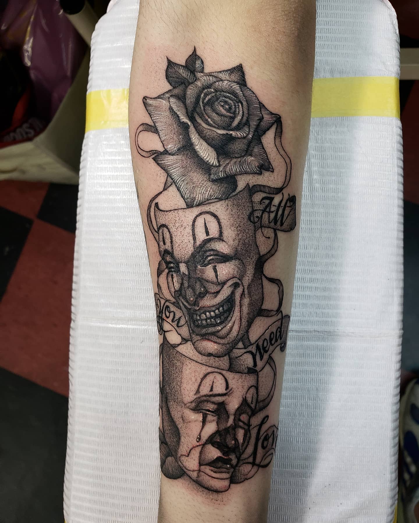 Tattoo of a rose, cheerful mask, and sad mask entwined with banner text on an inner forearm