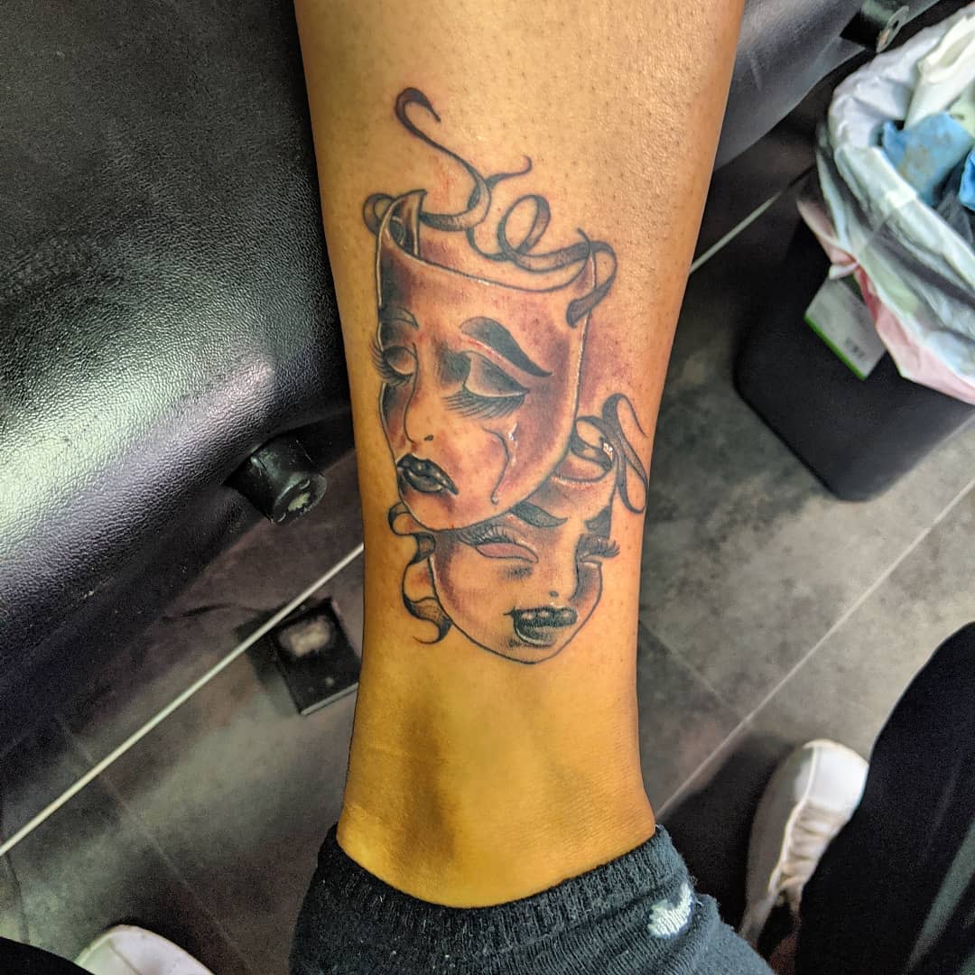 Forearm tattoo of contrasting drama masks depicting sorrow and joy with ribbon details