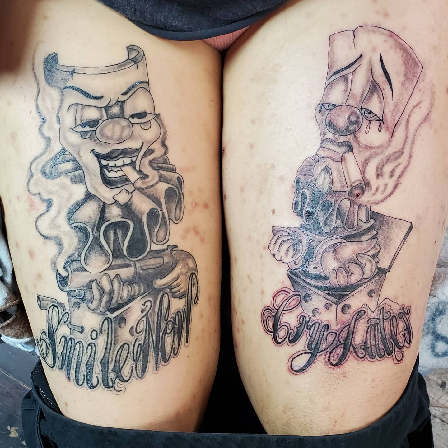 Close-up of tattoos on thighs depicting a laughing clown and a crying clown with text