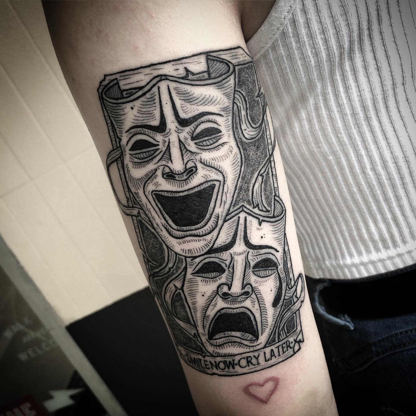 Tattoo on arm of comedy and tragedy masks above "Smile Now-Cry Later" text, heart below
