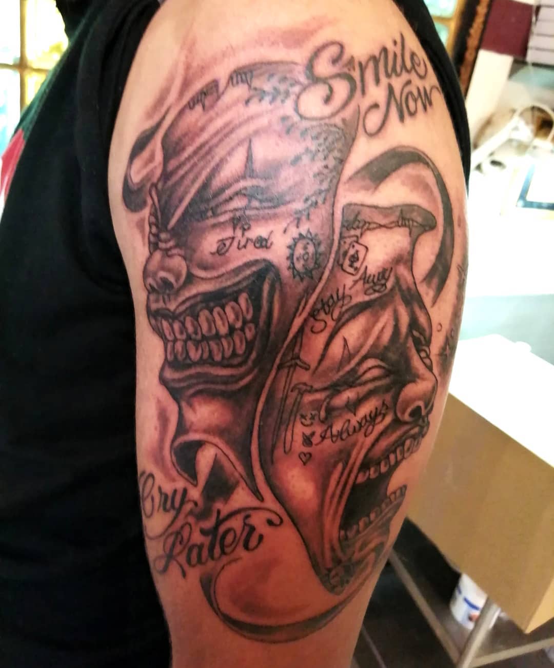 Upper arm tattoo of two masks with the text "Smile Now, Cry Later" and various detailed engravings