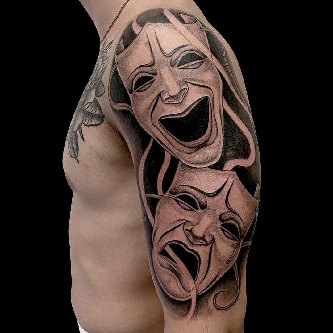 A bicep tattoo depicting comedy and tragedy masks in black ink shading