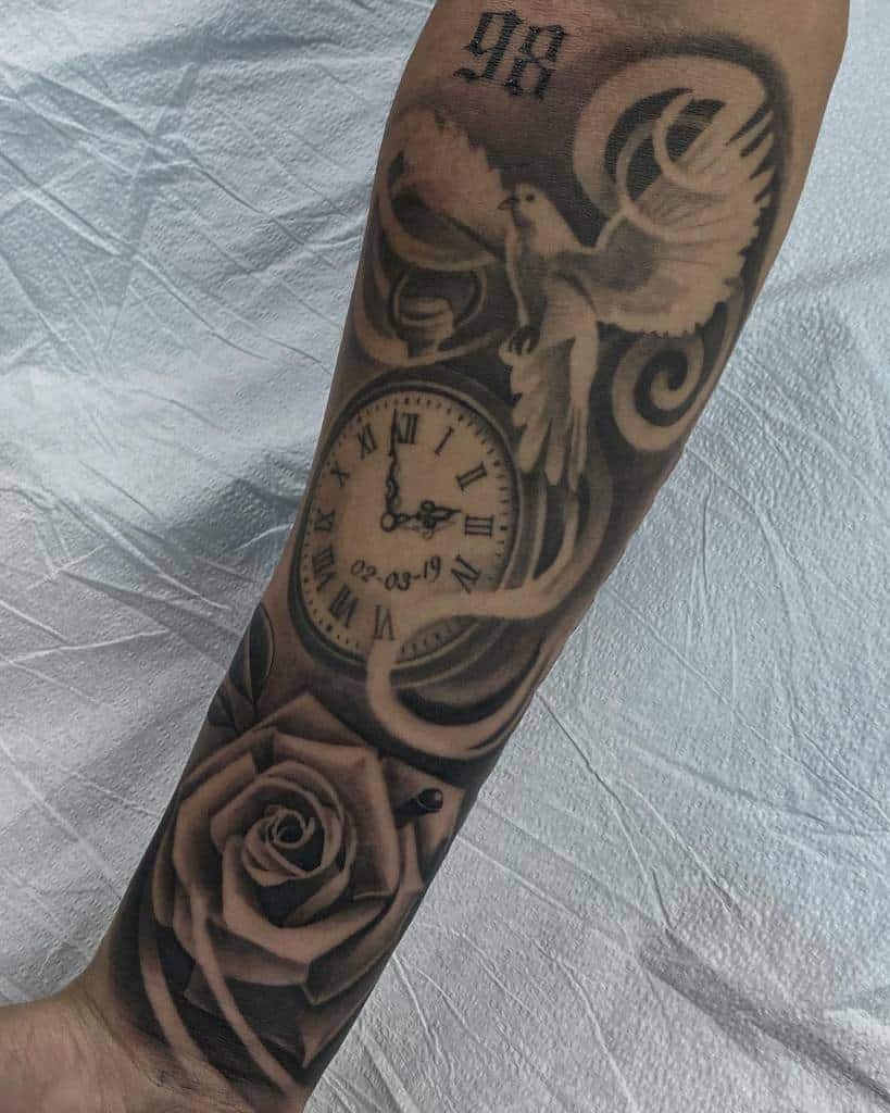 A black and grey forearm tattoo featuring a detailed dove in flight, a pocket watch with Roman numerals and a date, swirling smoke patterns, and a realistic rose