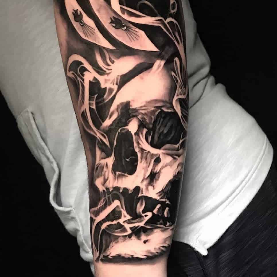 A black and grey forearm tattoo featuring a detailed skull enveloped in swirling smoke, creating a dramatic and eerie effect, with shading and contrast for depth