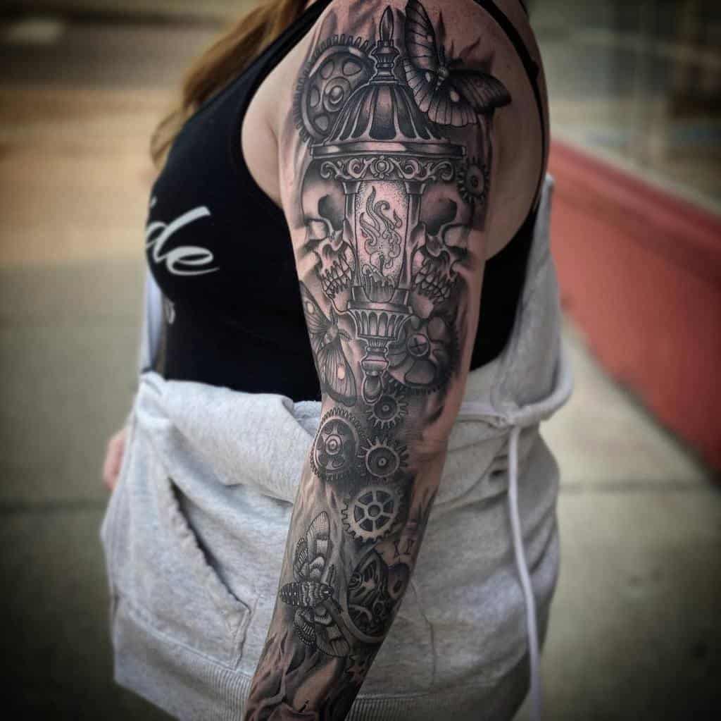 A detailed black and grey full sleeve tattoo featuring a steampunk-inspired design with gears, a lantern emitting smoke, and a butterfly, blending mechanical elements with soft shading for a dynamic look