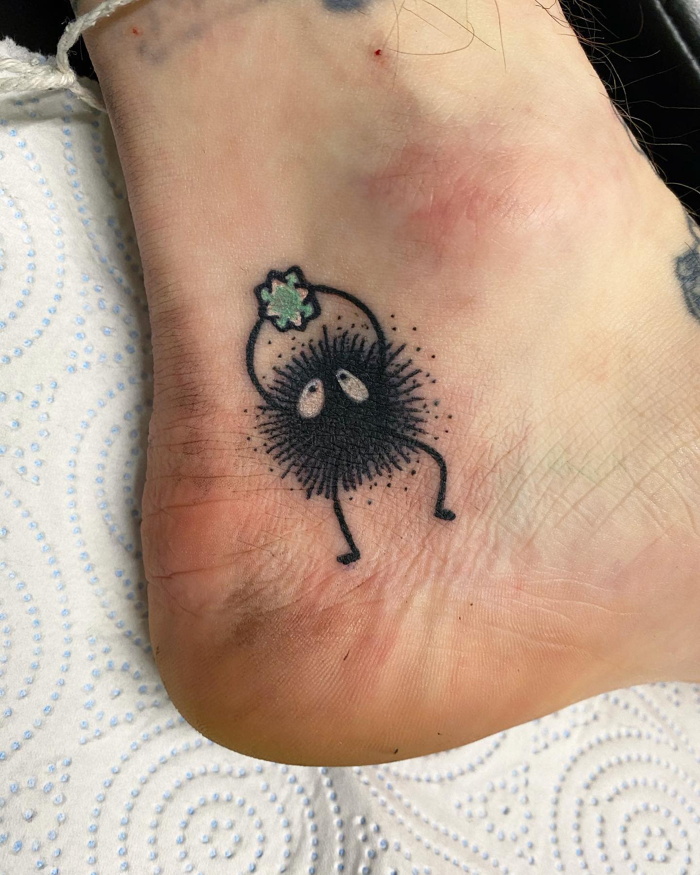 Simple black soot character tattoo with a green star atop its head on a foot
