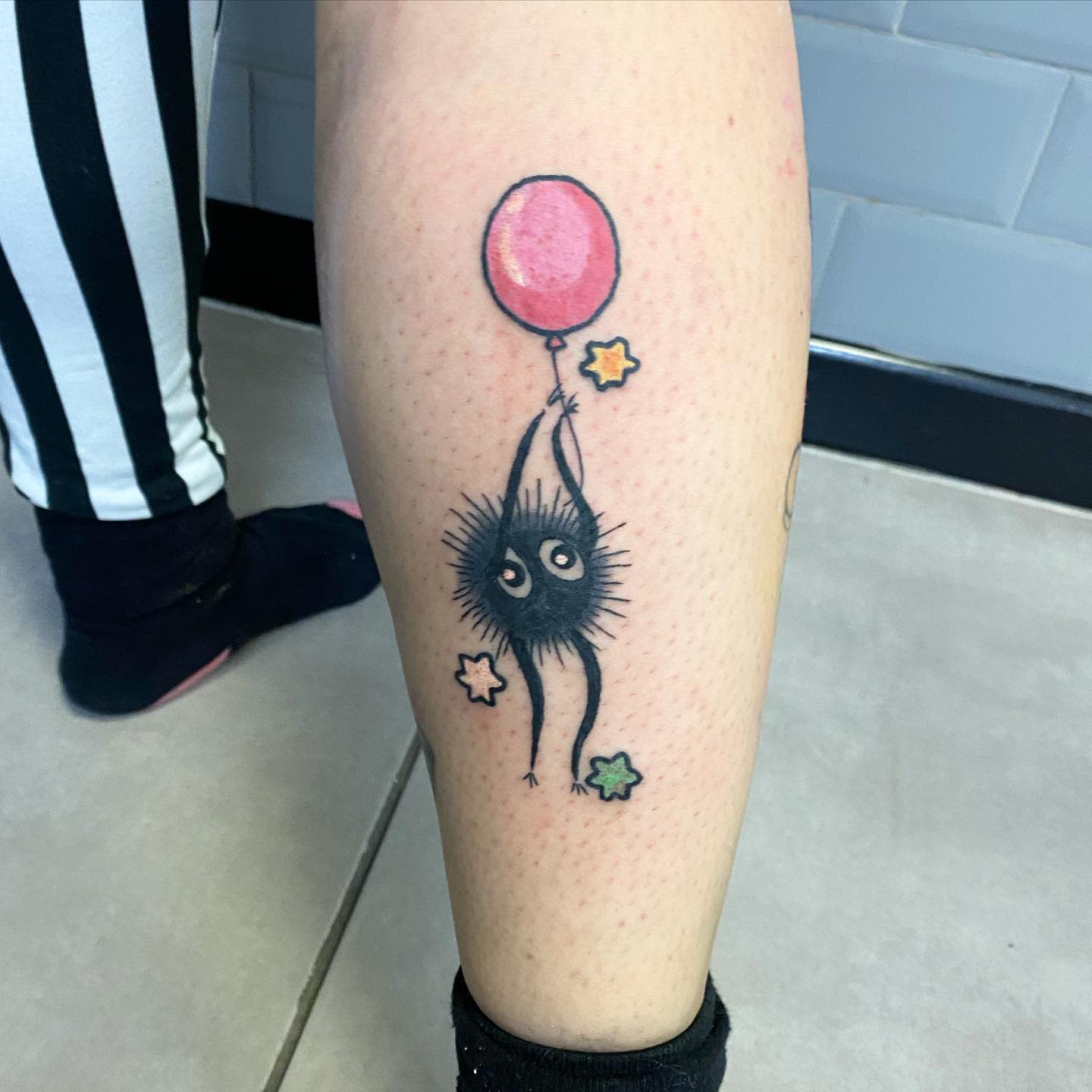 Tattoo of a soot sprite holding a pink balloon and colored stars on a leg