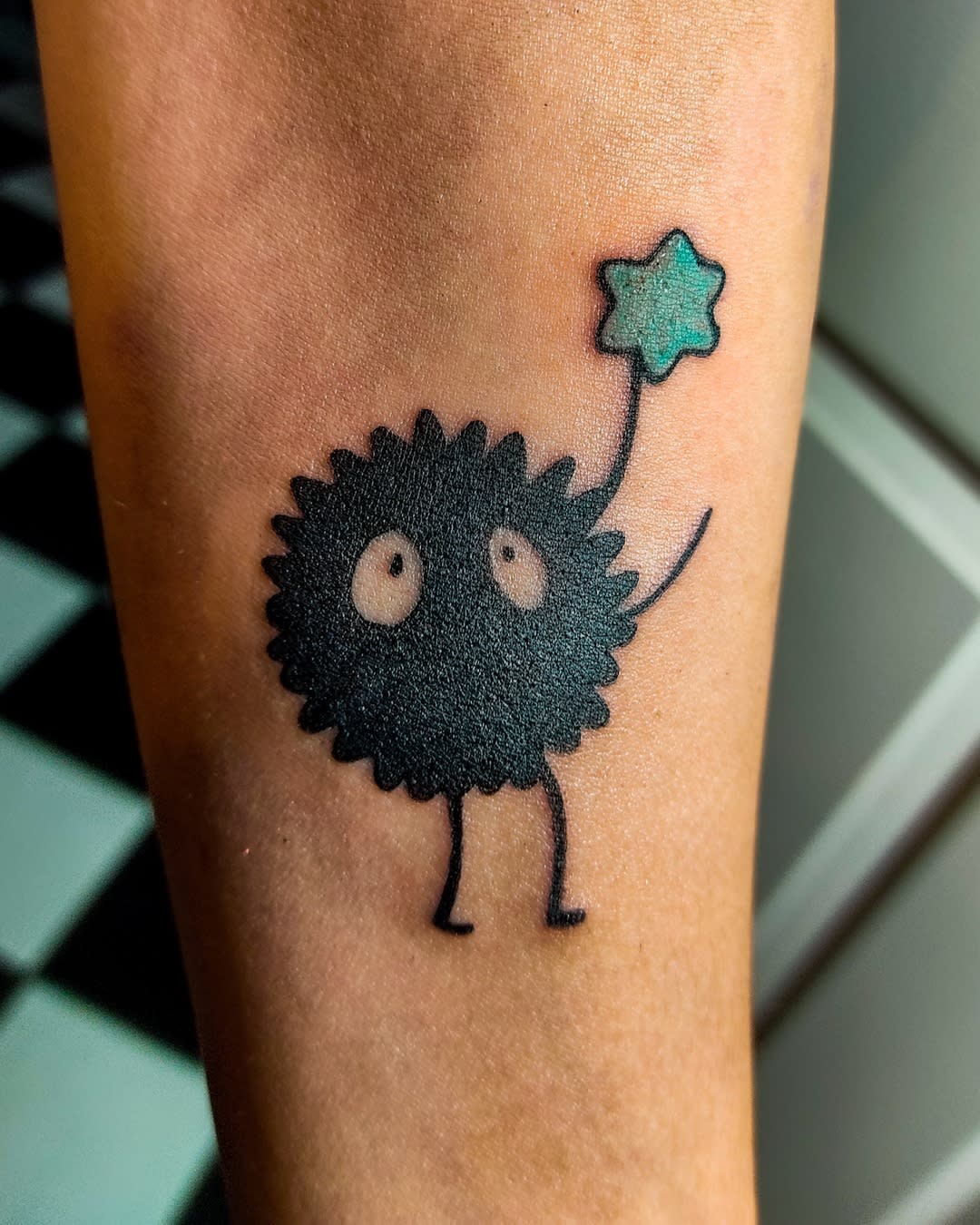 Tattoo of a soot sprite holding a green star on an arm