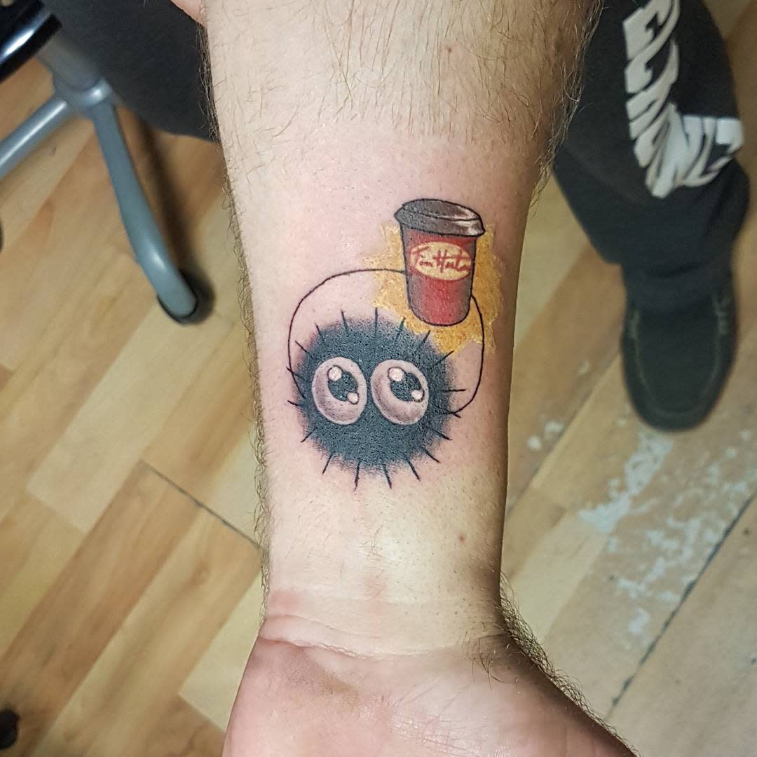 Cute soot sprite tattoo holding a coffee cup on a forearm