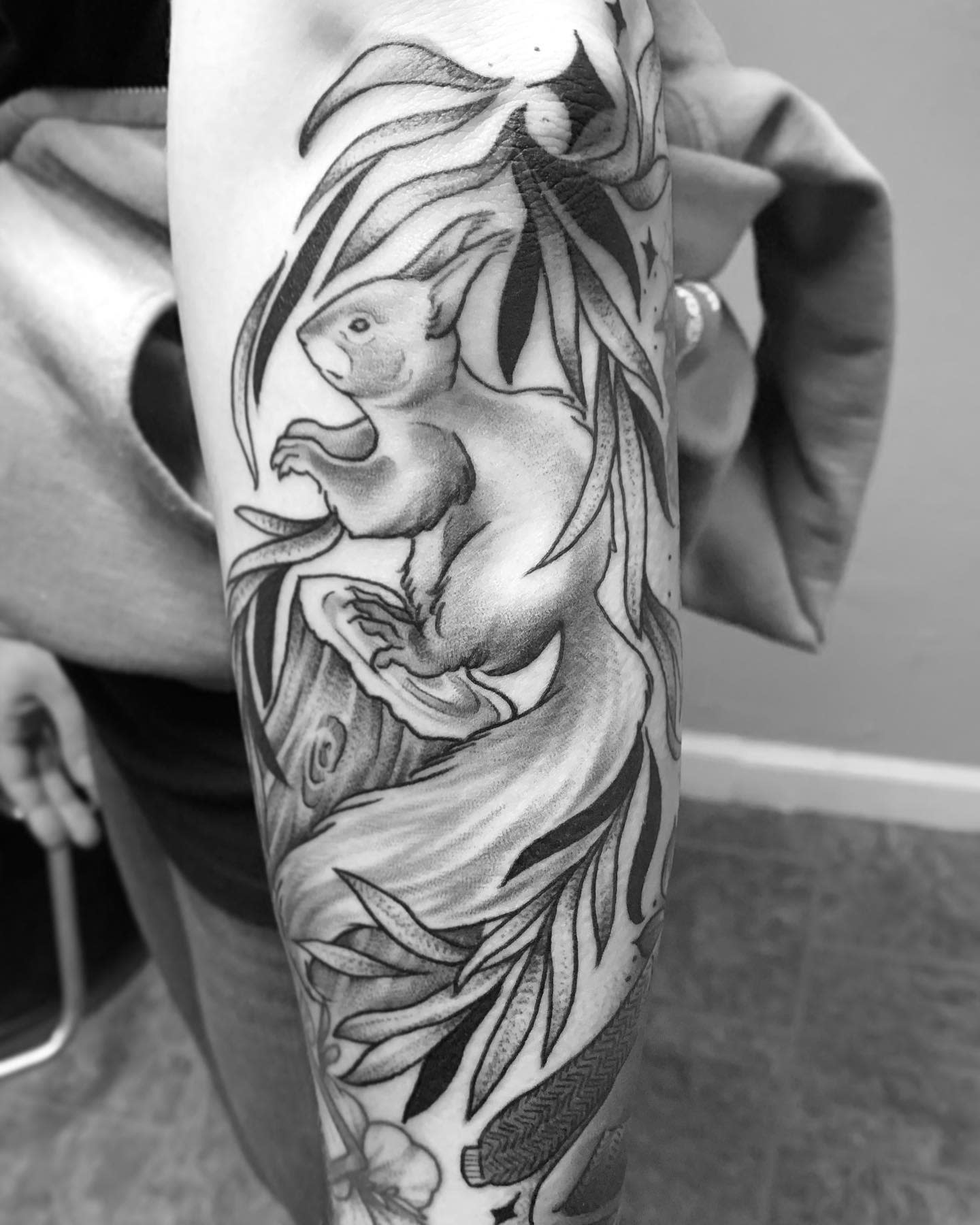 Black and white tattoo of a squirrel with leaves and branches on a forearm