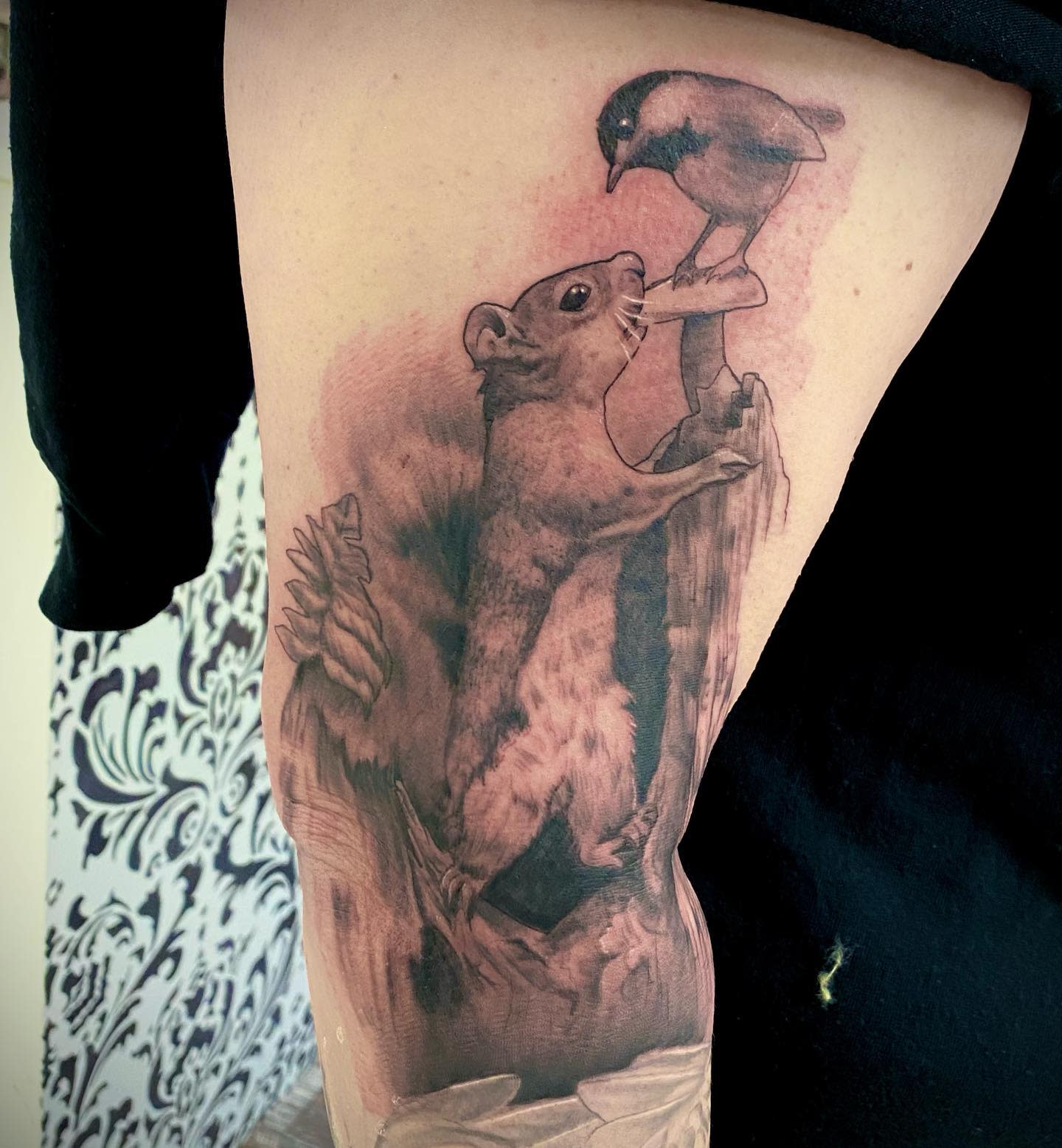 Tattoo of a squirrel climbing a branch to meet a bird on an upper arm