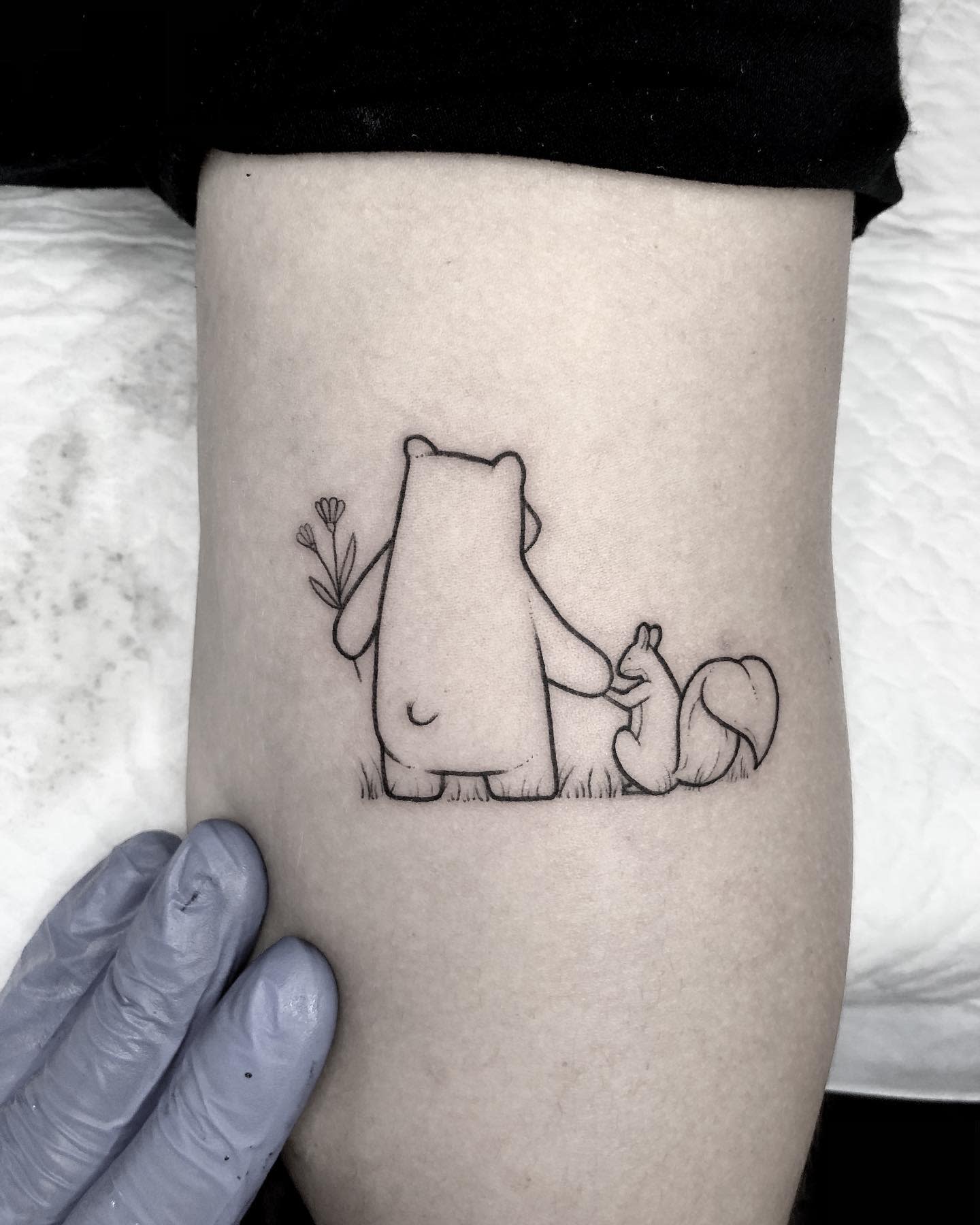 Outline tattoo of a bear holding flowers and a fox sitting beside it, on a leg
