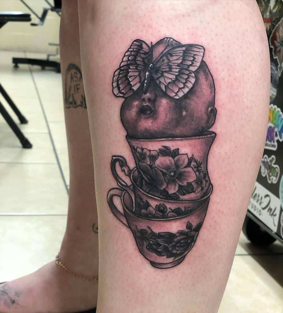 Black and grey tattoo of a baby doll head with a butterfly on top, stacked in three floral teacups