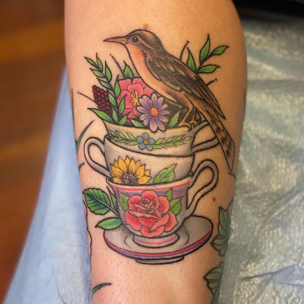 Colorful tattoo of three stacked teacups with floral patterns, the top cup filled with flowers, a bird perched on the rim, and a saucer at the bottom, creating a nature-inspired design