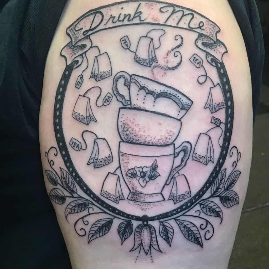 Black and grey tattoo of stacked teacups inside a circular frame with tea bags, swirling steam, leafy accents, and a 'Drink Me' banner