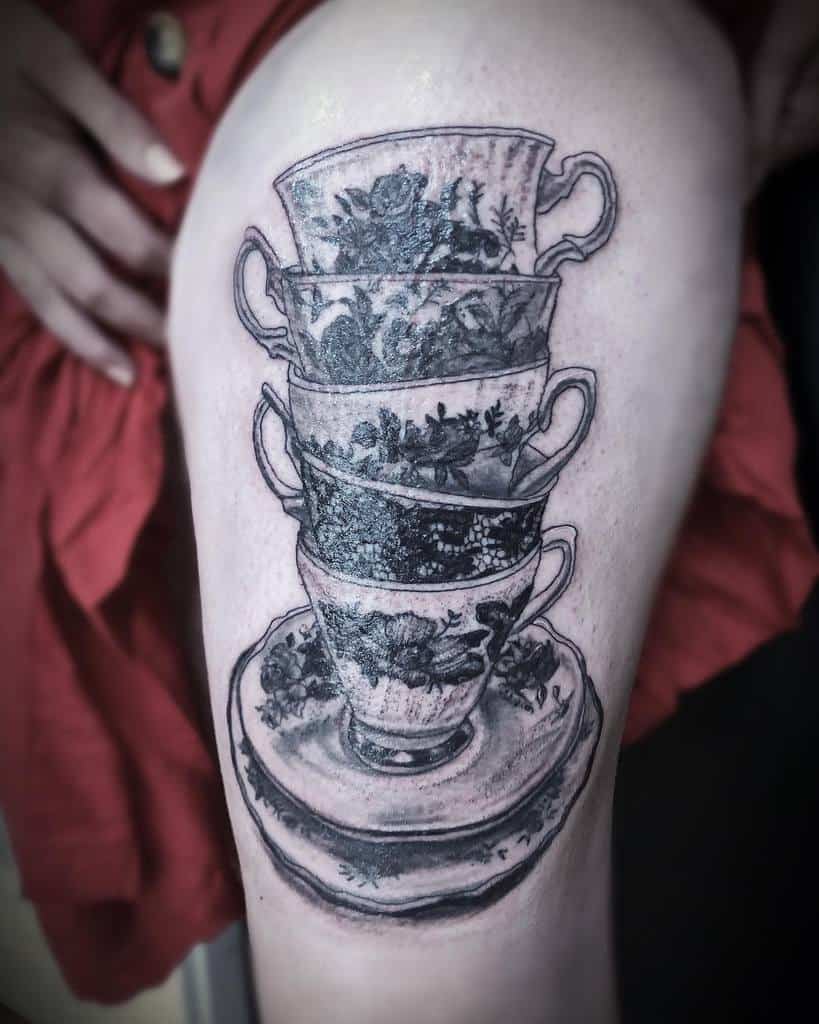 Black and grey tattoo of four stacked vintage teacups with intricate floral patterns, placed on two saucers for a classic and elegant design