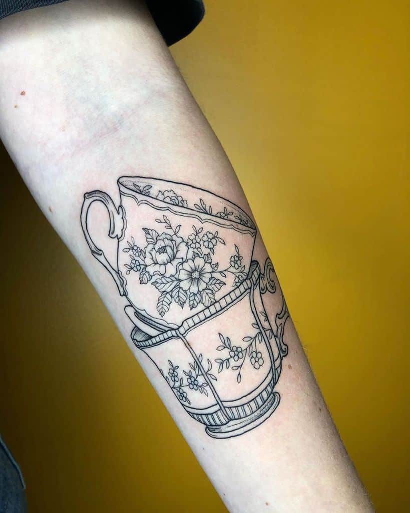 Black and grey tattoo of two intricately designed teacups with floral patterns stacked 